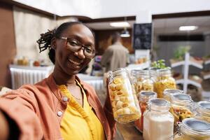 i bio-mat affär, svart kvinna bär glasögon innehar glas behållare lastad med spaghetti. organisk Produkter, vloggar, digital kamera, och kund videography är Allt exempel av uppkopplad marknadsföring. foto
