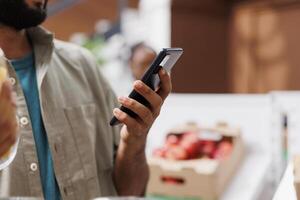 närbild på digital mobil enhet varelse hölls förbi en skäggig man i en eco vänlig bekvämlighet Lagra. selektiv fokus på kund använder sig av smartphone för granskning hållbar matvaror lista. foto