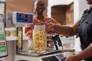 svart kvinna med glasögon väntar medan Säljare användningar digital skala till mäta glas behållare fylld med färsk pasta. afrikansk amerikan säljare vägning burk av nyligen tillverkad spaghetti för kund. foto