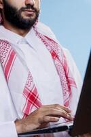 arab man i thobe och slöja innehav bärbar dator och skrivning meddelande på tangentbord närbild på hand. muslim person bär traditionell kläder använder sig av bärbar dator, textning och skriver foto