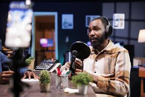 afrikansk amerikan influencer i de studio, använder sig av en avancerad mikrofon för ett internet program på en live streaming kanal. man håller på med en podcasting session med kvalitet strömning inspelning Utrustning. foto