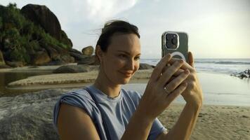 Söt ung caucasian kvinna tar bilder av de skön hav på tropisk ö strand. handling. Lycklig flicka på semester fotografier på smartphone med handflatan träd på de bakgrund. foto