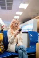 Lycklig asiatisk turist kvinna använder sig av mobil smartphone med resväska reser mellan väntar för flyg i flygplats terminal, flyg kolla upp i, turist resa resa begrepp foto