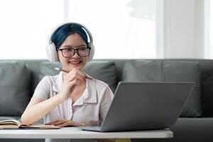 Lycklig ung kvinna Tonårs bär hörlurar skrivning notera. studerande uppkopplad inlärning klass studie uppkopplad video ring upp zoom lärare med bärbar dator och bok foto