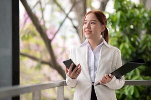 asiatisk företag kvinna skön med smart telefon och dokumentera fil, asiatisk kvinna med smartphone gående gå till arbete. förberedelse för de arbetssätt dag foto