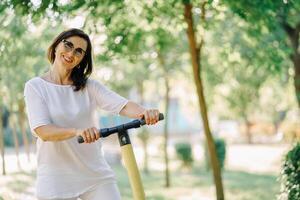 stänga upp glad förtjusande senior kvinna använder sig av en skoter medan ridning i de parkera. modern kvinna, en ny generation. friska glad senior pensionerad lady. begrepp av ålder inkludering och ekologisk foto