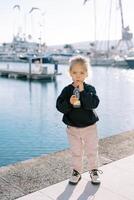 liten flicka drycker juice genom en sugrör från en flaska medan stående på de promenaden foto