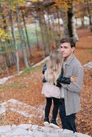 pappa kramar en liten flicka stående på en sten väg på en kulle i de höst skog och utseende in i de distans foto