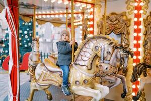 liten flicka rider en leksak häst på en karusell i de fyrkant nära en dekorerad jul träd foto