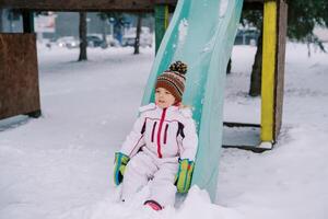 liten flicka glidning ner en snötäckt färgrik glida foto