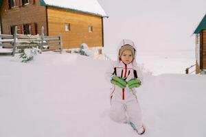liten flicka står i en djup snödriva nära trä- stugor foto