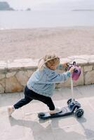 liten flicka rider en skoter längs en kaklade väg längs de stranden, tryckande av med henne fot foto