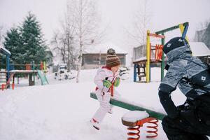 mamma och liten flicka rida på en snöig svängbalanserare på en färgrik lekplats foto