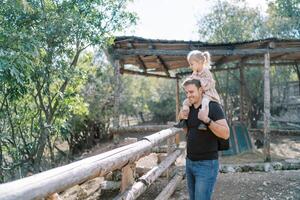 leende pappa med liten flicka på axlar stående nära trä- staket av inhägna i parkera foto