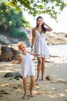 ung kvinna mor med en liten dotter i vit klänningar har en roligt på havsstrand i de skugga av träd och handflatorna. hög kvalitet Foto