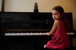 äkta skön liten barn flicka i elegant röd klänning, ser på kamera, Sammanträde på piano och utför klassisk melodi på stor piano. förtjusande barn flicka spelar piano på Hem. kopia Plats foto