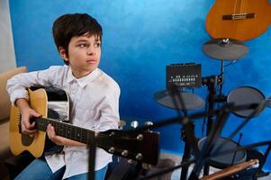 äkta porträtt av ett tonåring pojke spelar gitarr i de modern musik studio. elektrisk gitarr hängande på en blå vägg nära trummor. människor. musik lektion. barn utbildning och underhållning foto