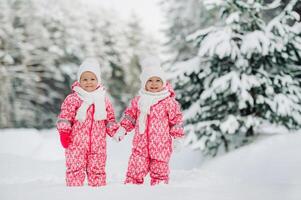 två liten tvilling flickor i röd kostymer stå i en snöig vinter- skog foto