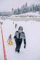 leende mor drar en kälke med en små barn upp en kulle på en åka skidor backe foto