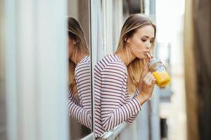 ung kvinna dricker ett glas naturlig apelsinjuice, lutad ut genom fönstret i hennes hem.