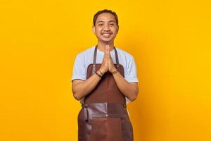 porträtt av leende stilig asiatisk ung man som bär förkläde och hälsar kunden med ett stort leende på läpparna isolerad på gul bakgrund foto