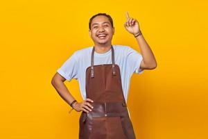 gladlynt stilig asiatisk ung man bär förkläde som pekar uppåt för att kopiera utrymme isolerat på gul bakgrund foto