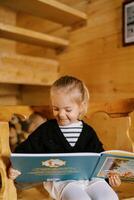 liten leende flicka läsning en färgrik bok medan Sammanträde på en trä- stol foto