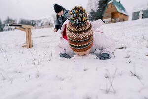 liten flicka lögner på henne mage på en snöig kulle nära henne Sammanträde mor och smakar de snö foto