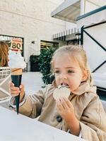 liten flicka äter en våffla från ett is grädde kon medan Sammanträde på de tabell foto