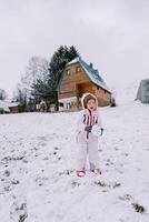 liten skrattande flicka med en snöboll i henne händer står på en snöig backe nära en trä- hus foto