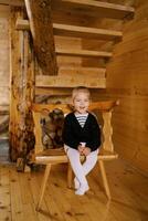 liten skrattande flicka Sammanträde på en stol nära de trappa i en trä- stuga foto