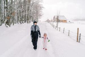 leende mor och liten flicka promenad längs en snöig by väg innehav händer foto