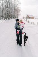 leende mor med en liten flicka i henne vapen promenader med henne hund längs en snöig by väg foto