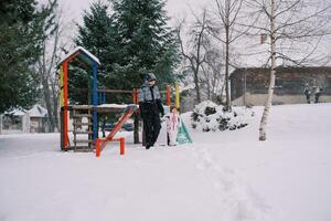 liten flicka och henne mor promenad innehav händer från en färgrik snötäckt glida genom de parkera under snöfall foto