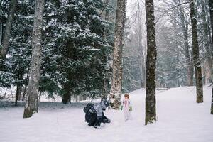 liten flicka står i en snöig skog och utseende på henne mamma framställning en snögubbe foto