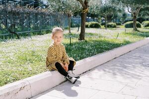liten leende flicka sitter på de trottoarkant nära en blommande äng foto