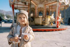 liten flicka med en teddy Björn står med en biljett i henne hand nära en spinning karusell foto
