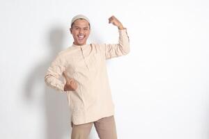 porträtt av religiös asiatisk muslim man i koko skjorta med kalott höjning hans näve, fira Framgång. prestation begrepp. isolerat bild på vit bakgrund foto