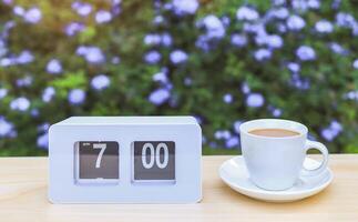 vit flip klocka som visar 7.00 o klocka och vit kopp av kaffe på de tabell i de trädgård med lila blommor bakgrund. foto