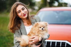 leende kvinna innehav en fluffig pomeranian hund nära en röd bil i de parkera foto