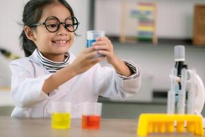 liten söt flicka med en mikroskop innehav en laboratorium flaska med vatten experimentera studie vetenskapsmän på skola. utbildning vetenskap begrepp. foto