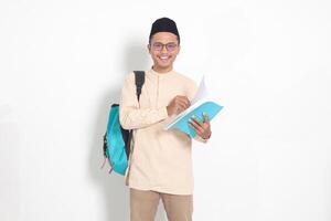 porträtt av upphetsad studerande asiatisk muslim man i koko skjorta med kalott bärande ryggsäck, medan innehav hans skola böcker. islamic utbildning begrepp. isolerat bild på vit bakgrund foto