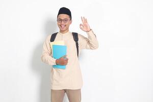 porträtt av upphetsad studerande asiatisk muslim man i koko skjorta med kalott bärande ryggsäck, innehav skola bok, som visar tumme upp gest. islamic utbildning begrepp. isolerat bild på vit bakgrund foto