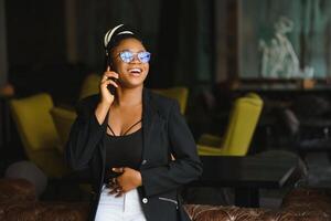 leende mörk flådd kvinna i trendig ha på sig lavning Kafé talande på mobil telefon via roaming tullar, afrikanska amerikan tusenåriga kvinna gående och framställning smartphone ring upp nöjd med förbindelse foto