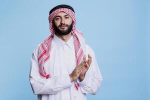 självsäker muslim man bär traditionell islamic kläder applåderar medan ser på kamera. ung arab klädd i vit thobe och rutig ghutra applåder händer studio porträtt foto