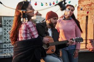 tre vänner njuter av att sjunga akustiska gitarrlåtar på taket foto