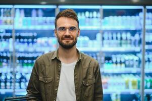 man med en skägg köper en öl i de alkohol avdelning av en mataffär . stilig man väljer en öl flaska i de Lagra och kommunicerar på en smartphone.shopping i en mataffär foto