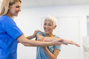 Lycklig senior kvinna håller på med övning med fysioterapeut. gammal pensionerad lady håller på med stretching vapen på klinik med de hjälp av en personlig tränare under en rehabilitering session. foto