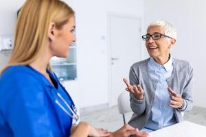 ung kvinna läkare eller gp i vit medicinsk enhetlig rådfråga kvinna patient i privat sjukhus. kvinna terapeut tala prata med senior kvinna klient på samråd i klinik. foto