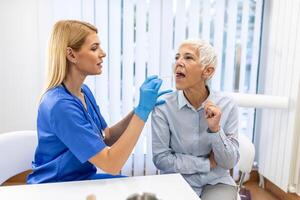 senior patient öppning henne mun för de läkare till se i henne hals. kvinna läkare granskning öm hals av patient i klinik. otolaryngologist undersöker öm hals av patient. foto